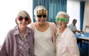 A group of three senior women with their arms around each other, smiling while they wear fun, silly glasses