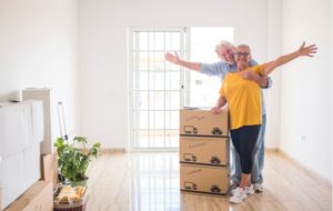 A senior couple holding their arms out in happiness beside three staked boxes as they are just moving into their new, more simplistic space in a senior community