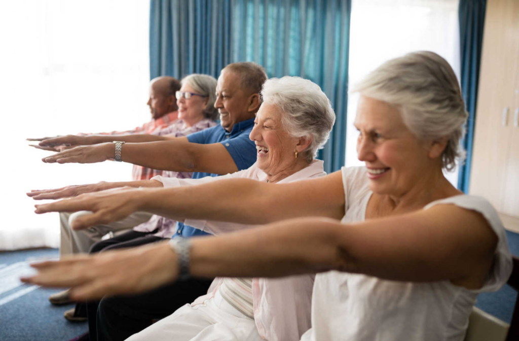 A group of senior living residents sitting in a row with their arms out in front of them as they participate in an exercise class.