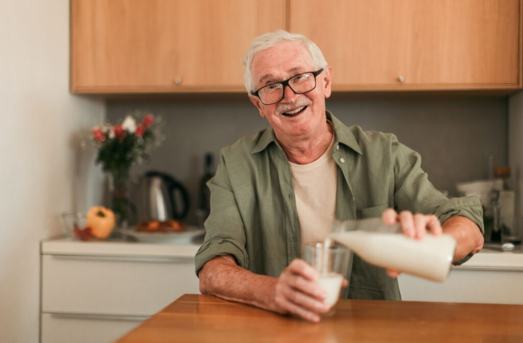 A senior man sitting in a kitchen and pouring milk in a glass