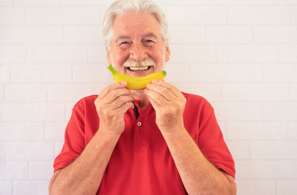 A senior man smiling with a white beard and in red polo shirt, holding a banana in front of his mouth like a smile