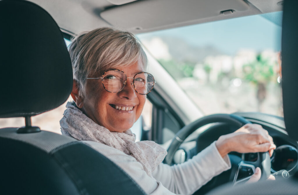 An older adult woman holding a car steering wheel, smiling and looking directly at the camera