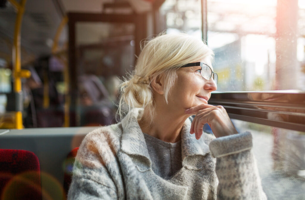 An older adult woman sitting on a public transport bus smiling and looking through the window