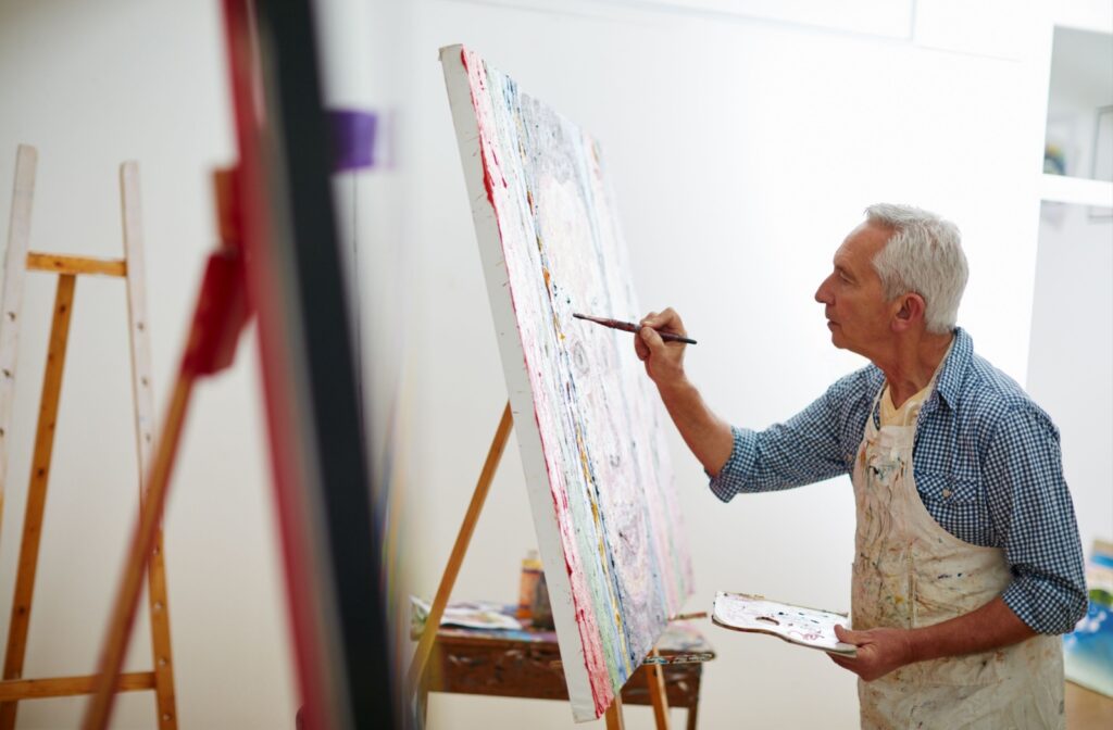 An older adult man holding a paintbrush and painting at home.