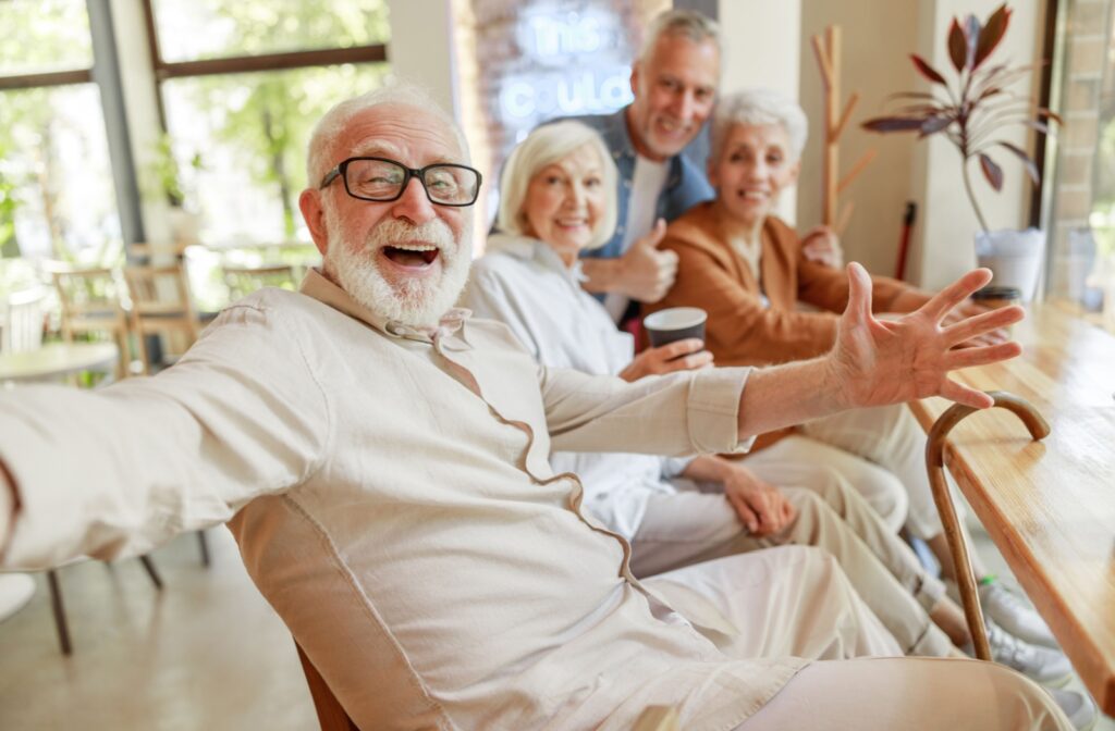 An older adult sits at a table with arms extended as if to give a hug and smiles at the camera.