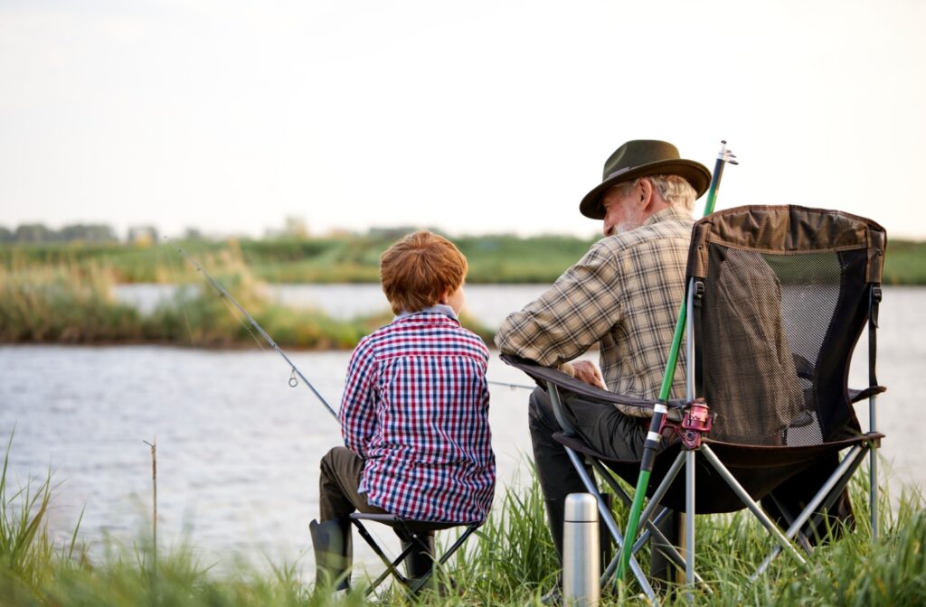 A senior and their grandchild sit side by side in lawn chairs. They chat and case fishing lines into the river in front of them.