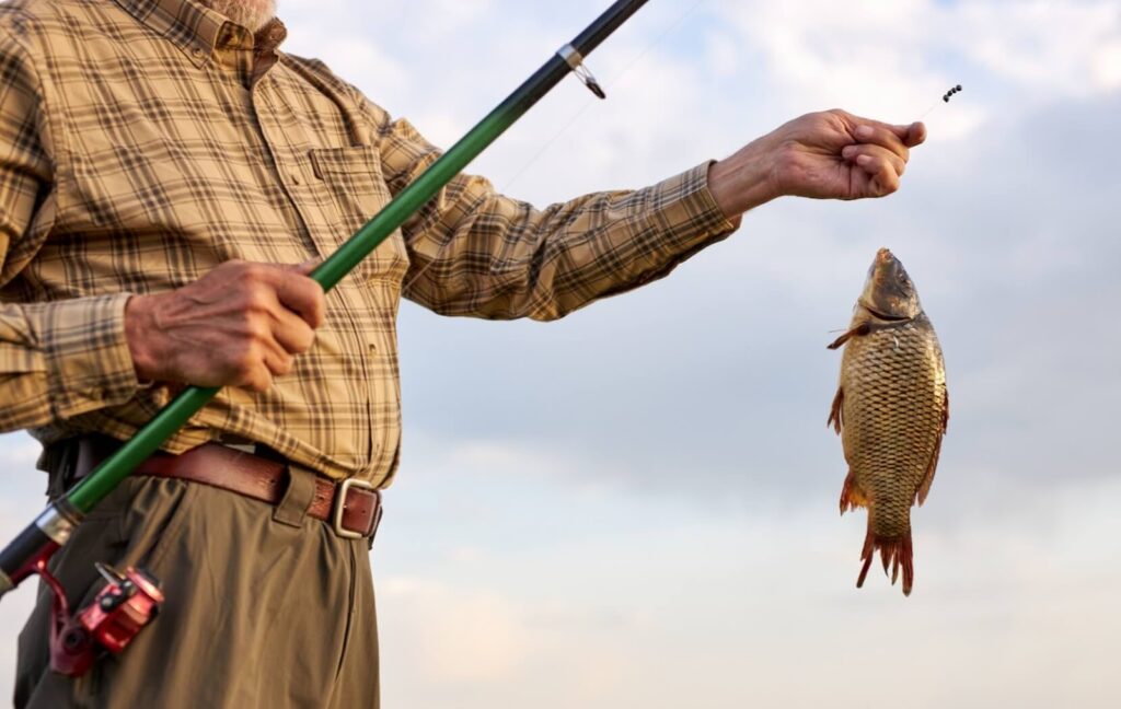 A senior wearing a plaid shirt pulls a small fish out of the water with their fishing rod during a quiet fishing trip.