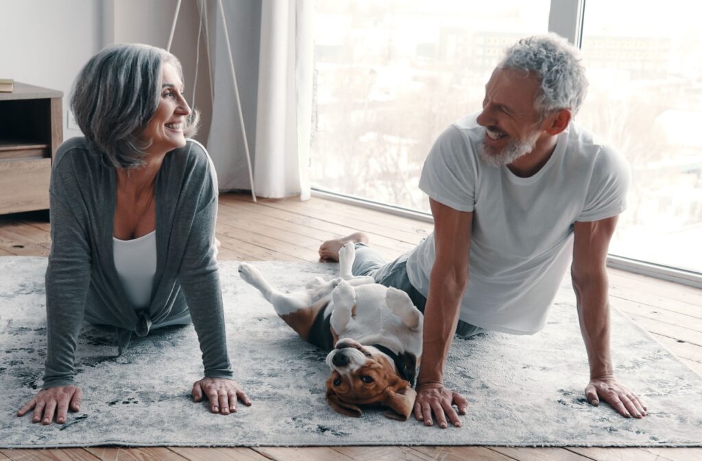 Older adults doing yoga on a rug indoors smiling at each other while a beagle lies happily between them with paws in the air
