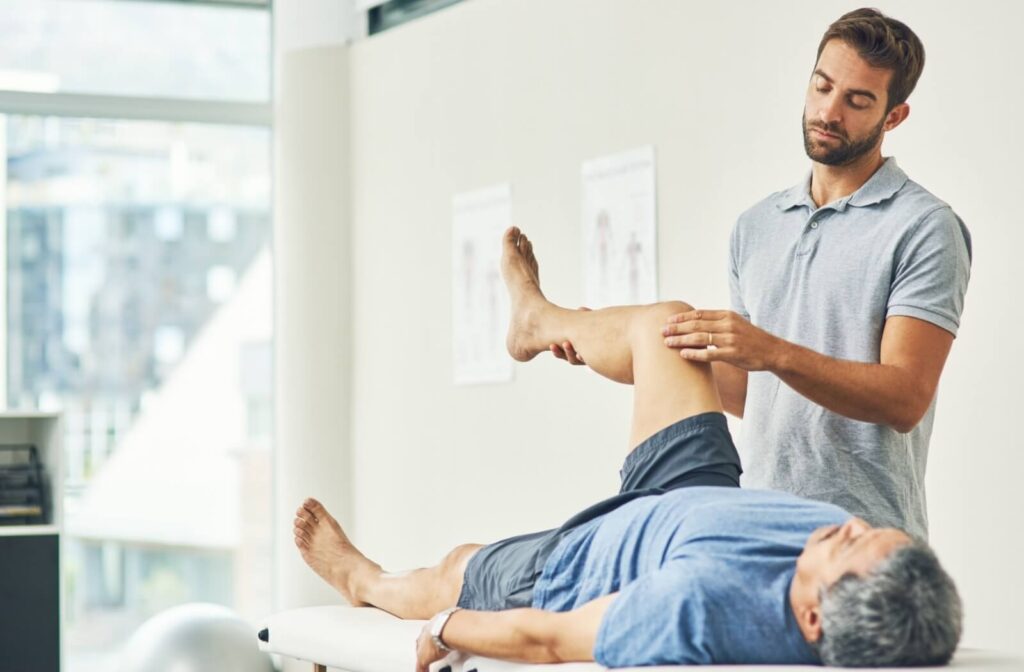 An older adult lies on their back while a physical therapist lifts their left leg to help them strengthen their knee