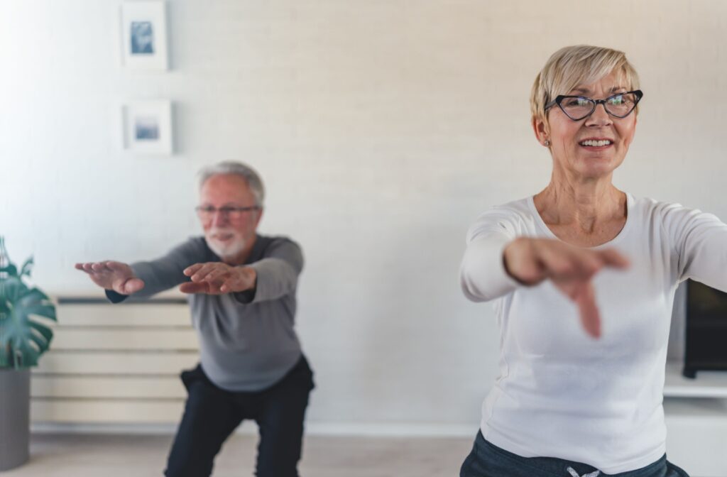 An older adult and their spouse smile as they perform standing squats in their living room to strengthen their knees